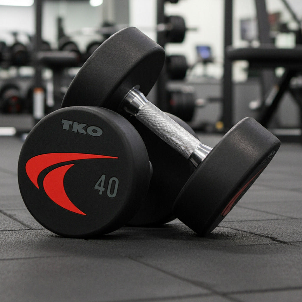 Pair of TKO 830US Signature Urethane Dumbbells shown in a gym setting.
