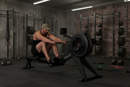 Man using the TKO 8AR AirRaid Rower in a gym setting with brick walls and various equipment.